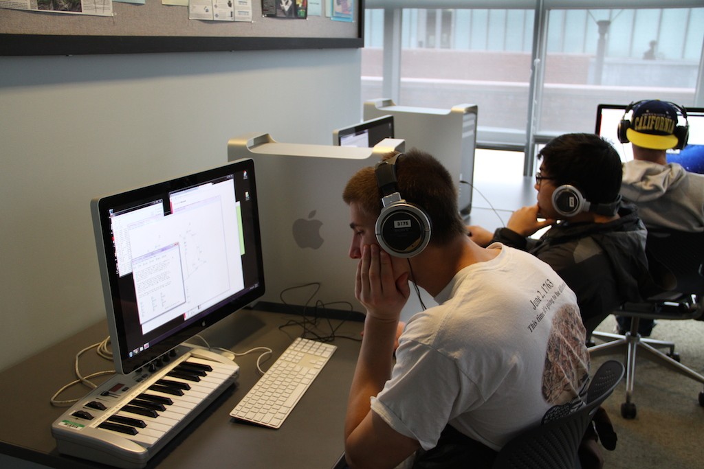 Two students wear headphones while working on laptops independently with an electronic keyboard sitting in front of one of them.