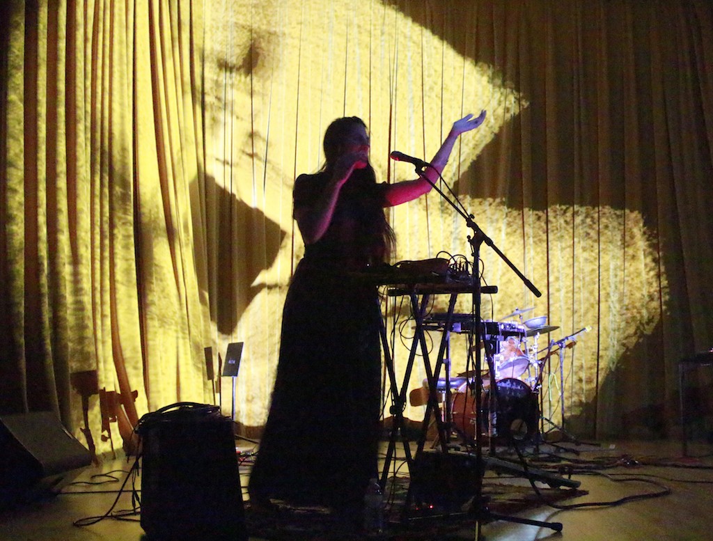 A women in dark silhouette, sings into a microphone with keyboard and electronics beside her on stage.