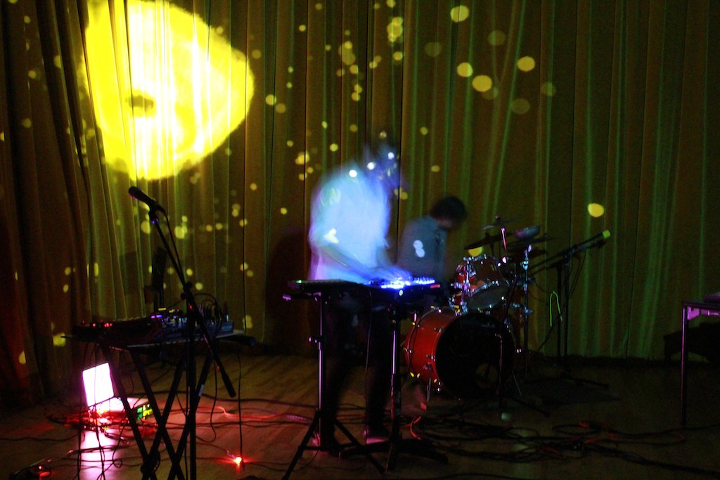 A blurred image of a person, performing on keyboard with electronics beside them and a drummer on stage.