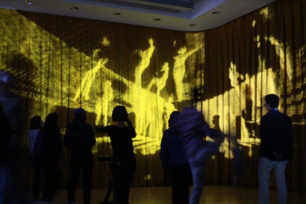 In a dark lit room, the silhouette of people dancing in front of a stage. 