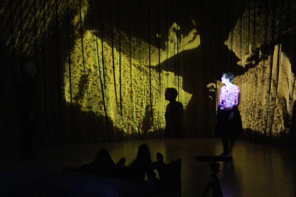 Two people stand in front of a yellow-lit backdrop casting large and oddly shaped shadows.