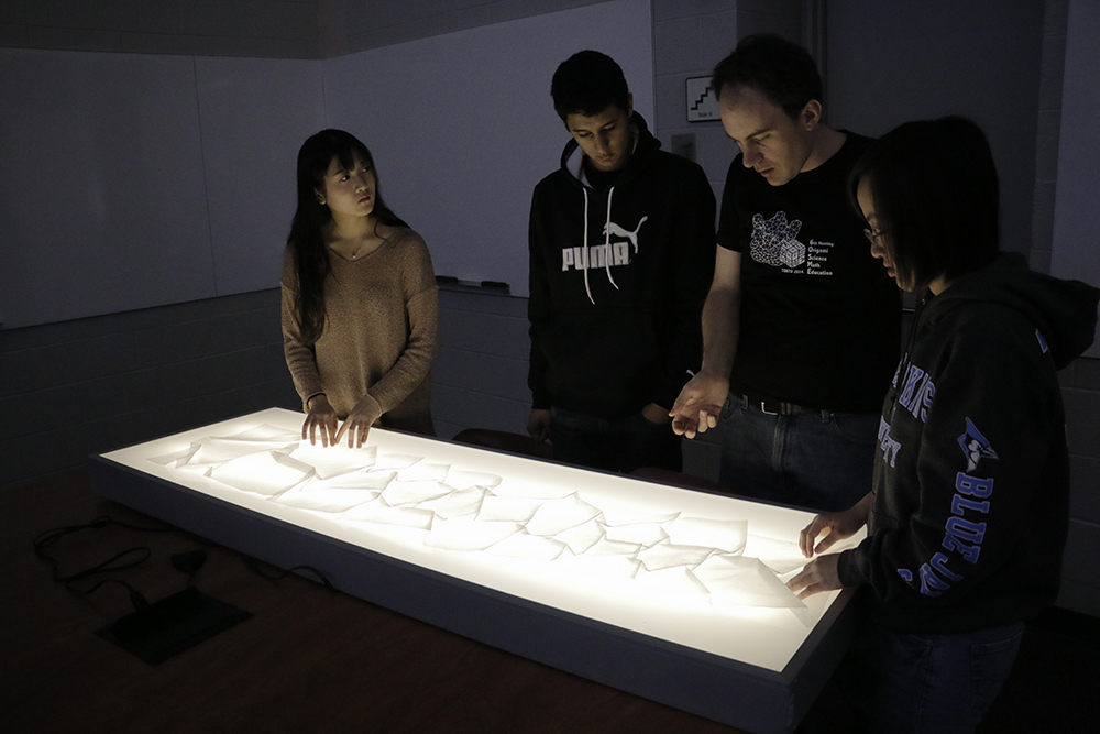 A group of students look at and discuss their work of a cosmic web made of glassine paper.