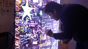 A student writes on an acrylic panel covered in neon writing.
