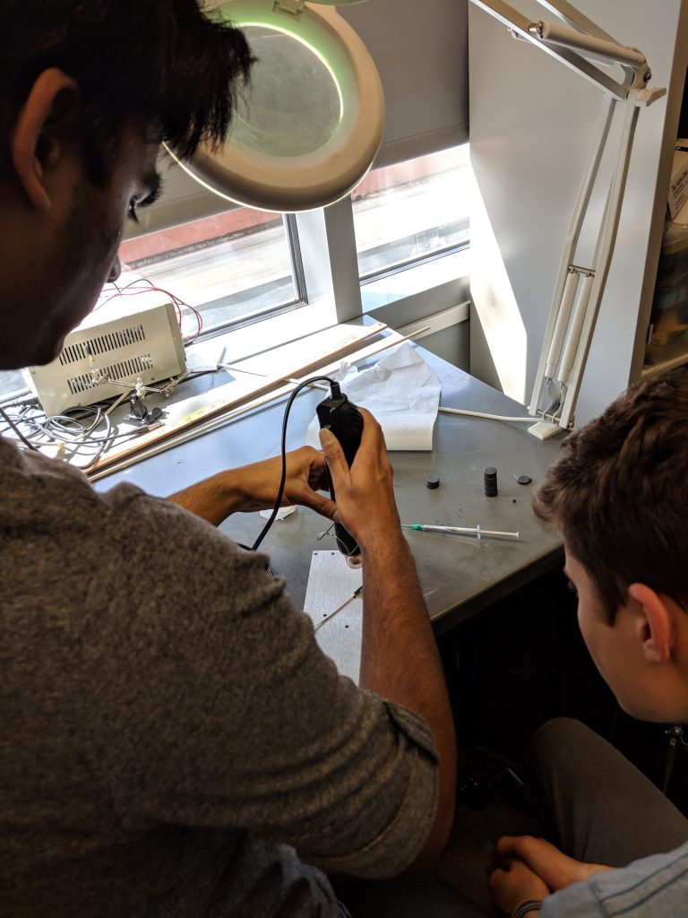 Standing behind two students, a guy uses a hand tool on a small piece of metal.