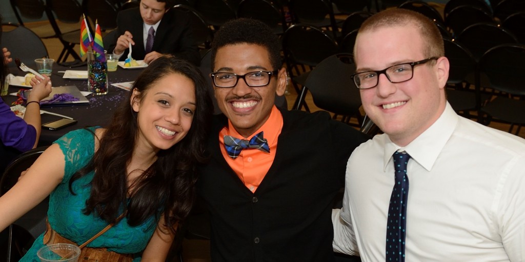 Three well dressed students get close together, with big smiles, while sitting at a table.
