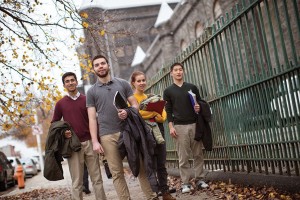 A group of students walk down the city sidewalk past a tall iron fence with fall colored leaves on the ground.