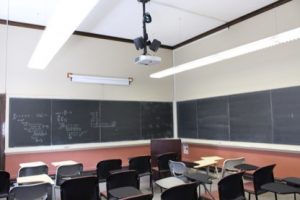 Empty classroom showing individual desk and chair seating with large black chalkboards on two sides of the room.
