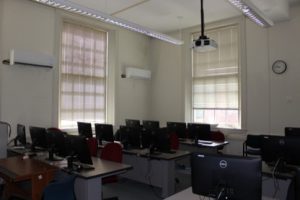Empty classroom showing tables and chairs with computer monitors at each seat and two large windows in the room.