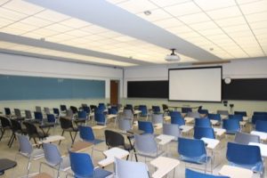 View of an empty classroom and individual student seating and desks and a large screen in the front of the room.