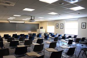 Empty classroom showing individual desk and chair seating; two windows on the right side. 