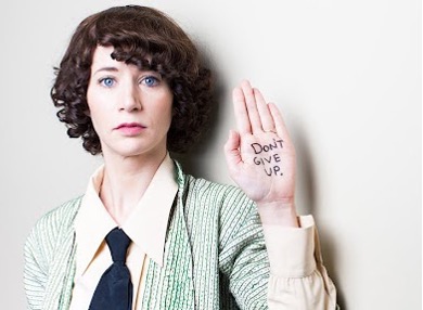 Miranda July holding up hand with writing "Don't Give Up".