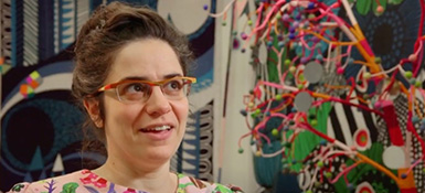 A photo of a woman in glasses speaking in front of a multicolored art project
