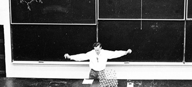A black and white photo of a man standing in front of a chalkboard in a lecture hall with his arms outstretched