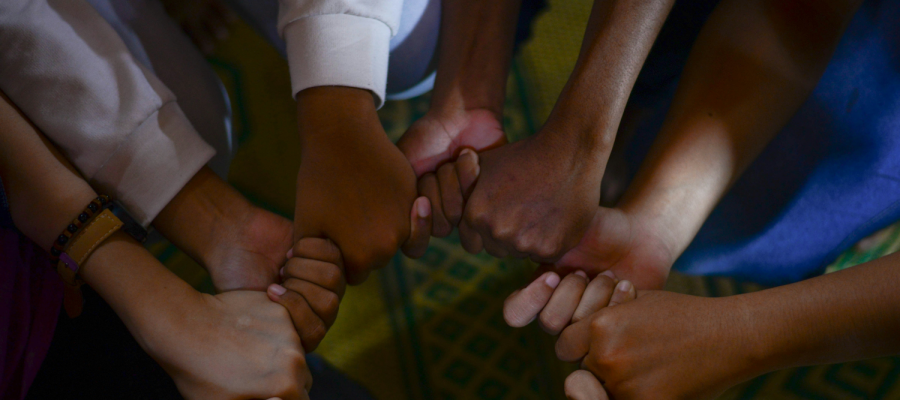 Full color image of various hands clasped in a circular configuration, implying unity and shared purpose.