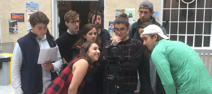 Multiple students stand behind a digital camera on a tripod, looking intently at the camera.