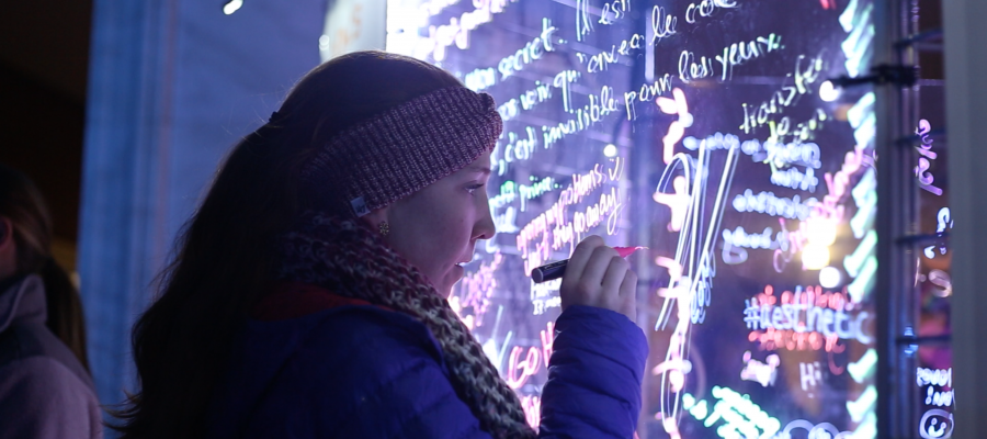 A student in a coat, scarf, and hat writes on an illuminated glass with glow in the dark ink.