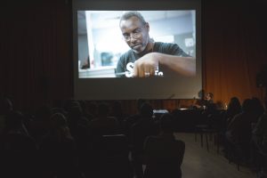 A dimly lit room with an image of an adult male on the large movie screen.