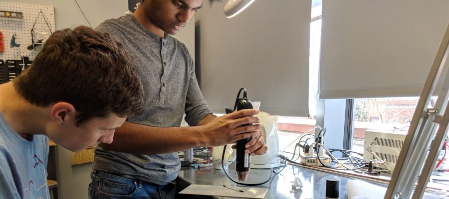 A side view of two students, one is using a hand tool on a small piece of metal.