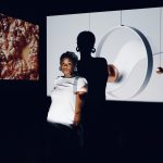 A woman stands in front of a light source casting her shadow on the screen behind her.