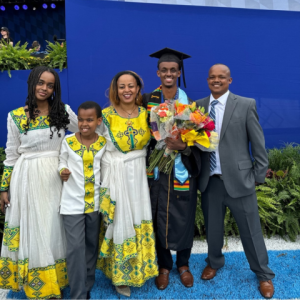 Family photo of Hopkins student Zerubabel Kebede. They are at commencement and he is wearing his graduation robe and cap. The family is smiling in front of a large blue screen.