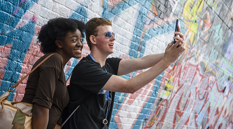Two students pose for a selfie in front of a multicolored graffiti wall