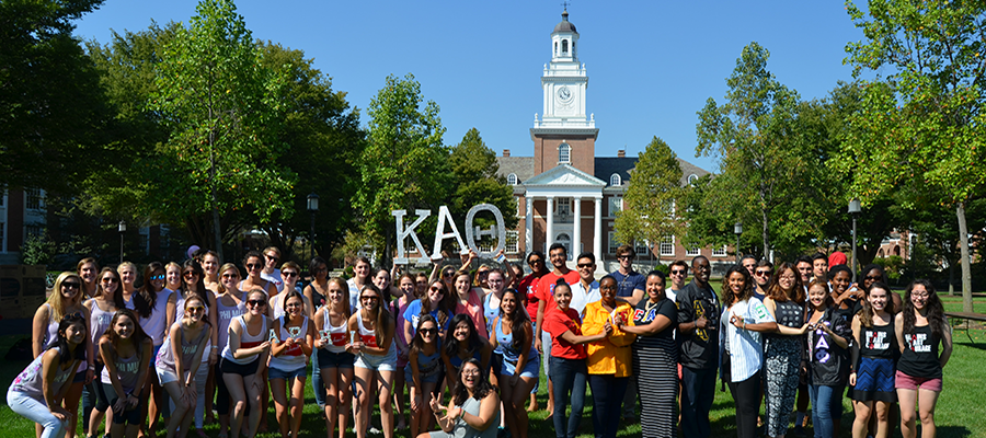 A large group of students pose for a photo while holding Greek letters in front of Gilman Hall