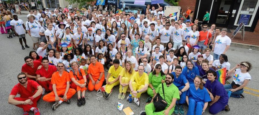 A large group of people sitting together wearing matching Pride shirts while a few individuals in the foreground wearing solid color scrubs that as a group depict the rainbow colors.