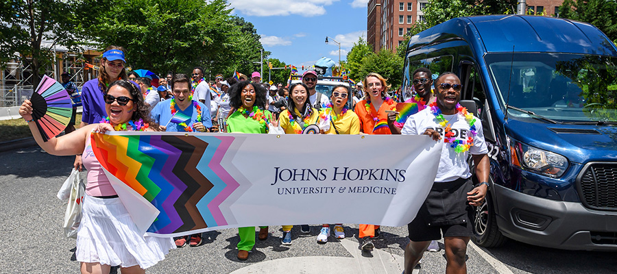 A group of people waving rainbow-colored flags and holding a banner that reads "Johns Hopkins Universiy and Medicine"