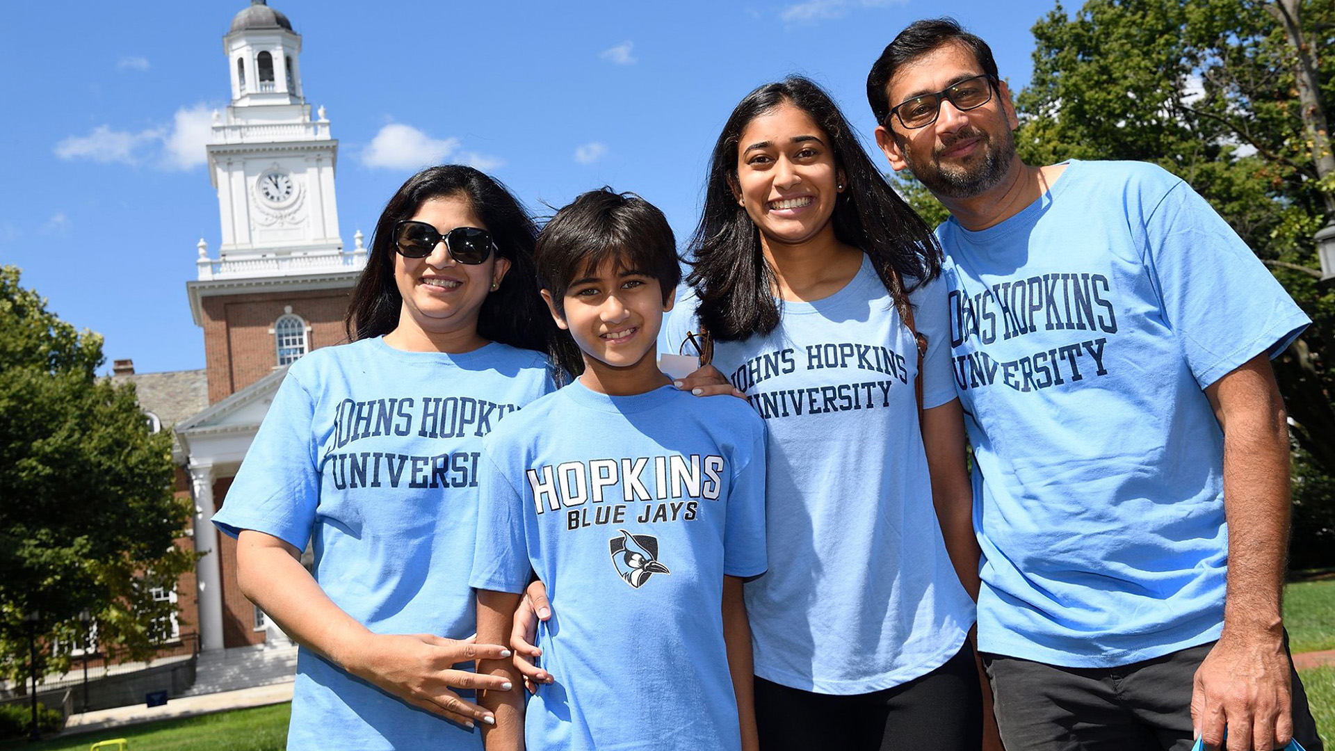 A family of four poses for a photo on Homewood campus while wearing Johns Hopkins t-shirts
