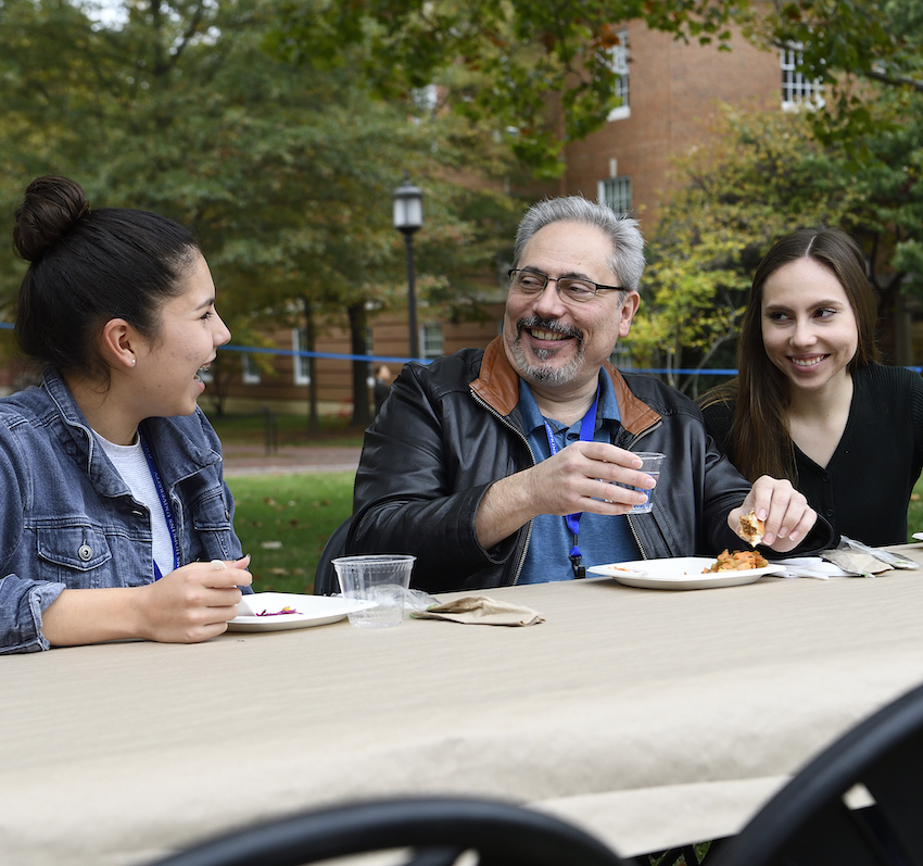 Two students smile while talking with their father at a table outdoors on campus