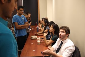 A group of students sit on one side of a table and talk with students individually on the other side.