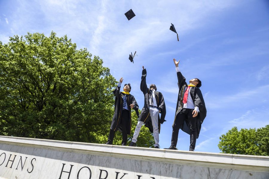 3 students stand on top of the Hopkins sign and throw their graduation caps.