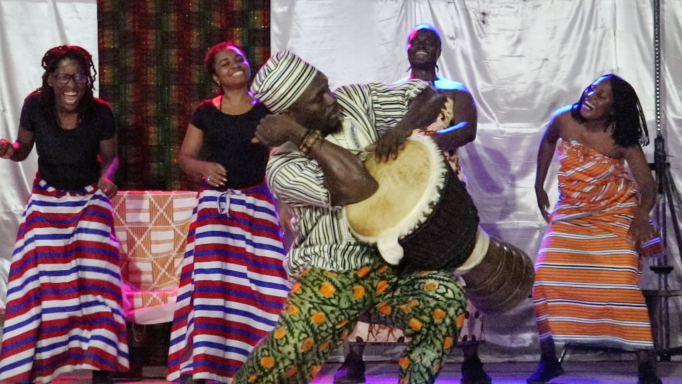 The performance, 200 Years of Returns, mid performance showing a man beating a drum with four women in the background smiling and dancing. Done through previous work by Dr. Jasmine Blanks Jones.