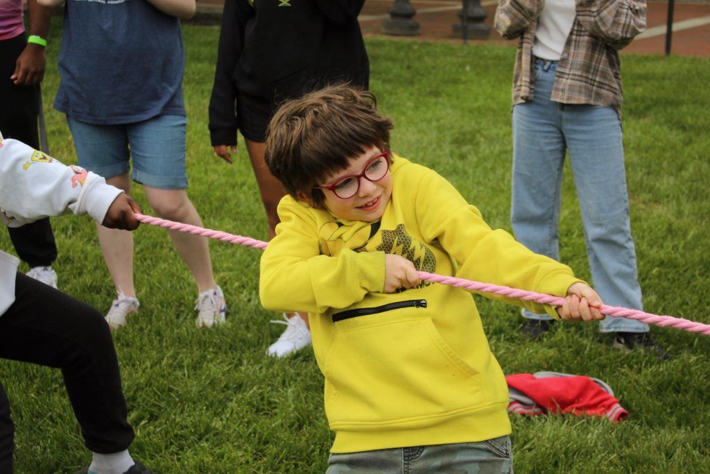 Tutee in a pikachu sweater smiling pulling on the rope in a tug of war game at the 2023 field day