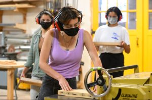 Frost working with tools. She's wearing a Lavendar tank top. There are two people in the back watching her as she cuts wood. This is in part of the CIIP program.