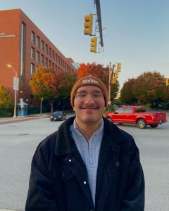 Hadeed Habib smiling. He is wearing a black jacket over a blue shirt. He has a orange-brown beanie on that says Modelo. He is standing at an intersection with a brick building in the background and a red truck behind him.