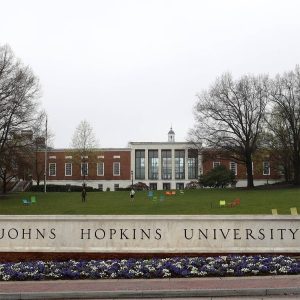 A photo of a marble sign reading "Johns Hopkins University" at the bottom of a grassy knoll on the Homewood campus. The MSE Library is in the background; the sky is cloudy.