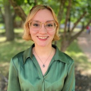 Eileen is smiling outdoors. She is wearing a green silky shirt and has glasses on.