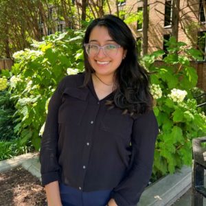 Headshot of Taz smiling outdoors in front of a green plant. She is wearing a dark long-sleeve button up and has glasses on. 