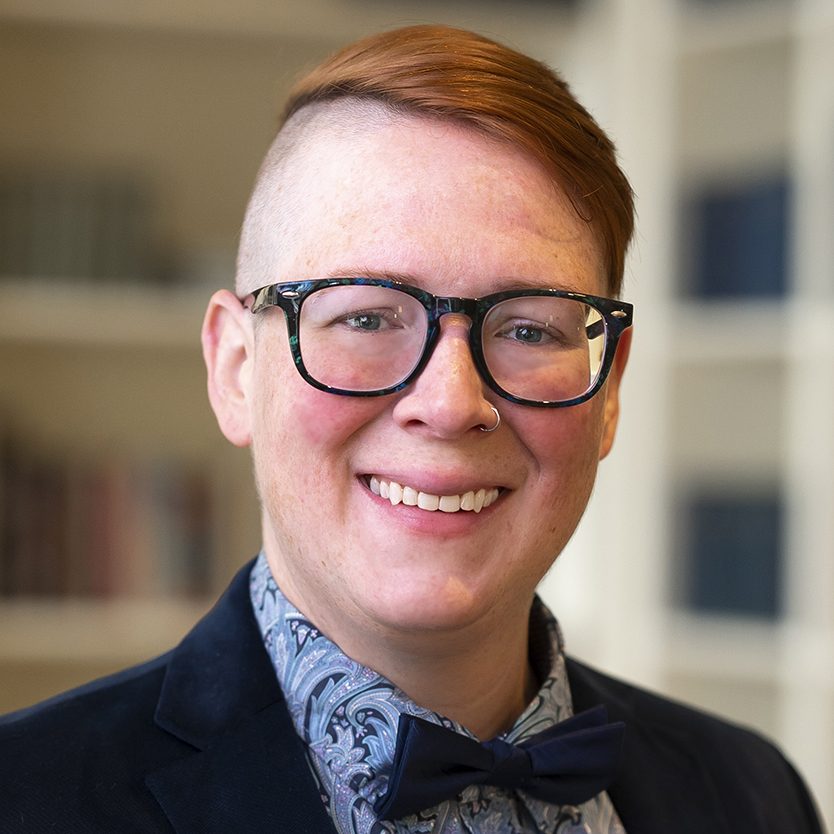 photo of sage magness-hill, a white, non-binary, young person with red hair and glasses. Sage is wearing a blue blazer, paisley print shirt, and navy bow tie. They are turned slightly to the right side of the photo, looking at the camera, and smiling.