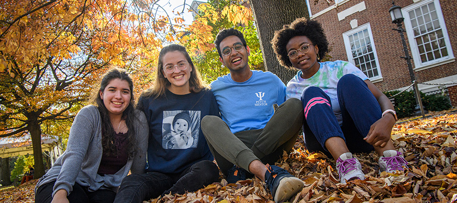 Four students sit together under a tree and smile, surrounded by autumn leaves