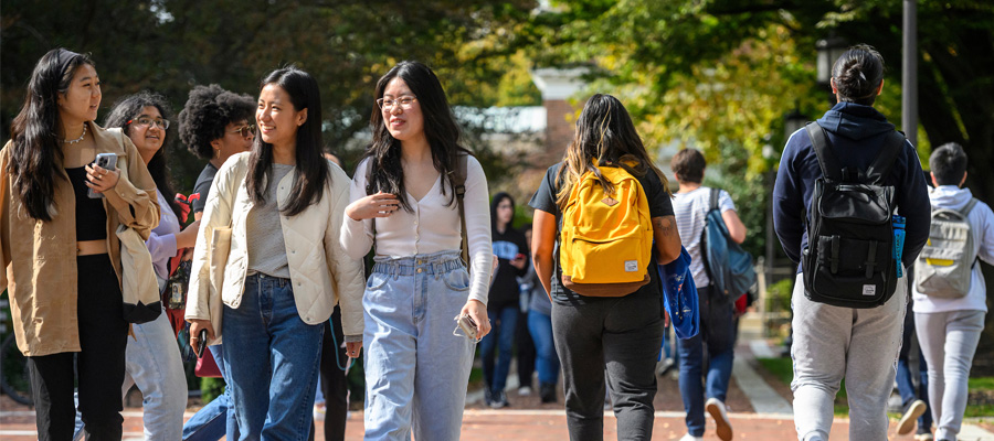 Three students talking and laughing with each other as they walk on campus. Behind them, other students, wearing backpacks, walk in various directions.